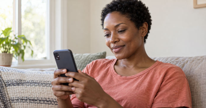 A woman in a coral shirt looking at a smartphone while seated on a sofa beside a bright home window.